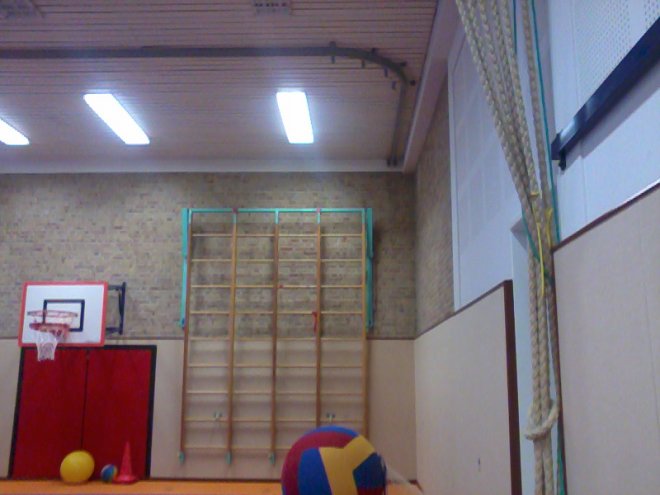 De gymzaal. Je ziet de touwenrij die gemakkelijk de zaal in en uit geschoven kon worden. Je ziet nog 1 wandrek. Het andere is verwijderd vanwege de nooduitgang. Je ziet ook nog een rail waaraan vroeger de ringen hebben gehangen.